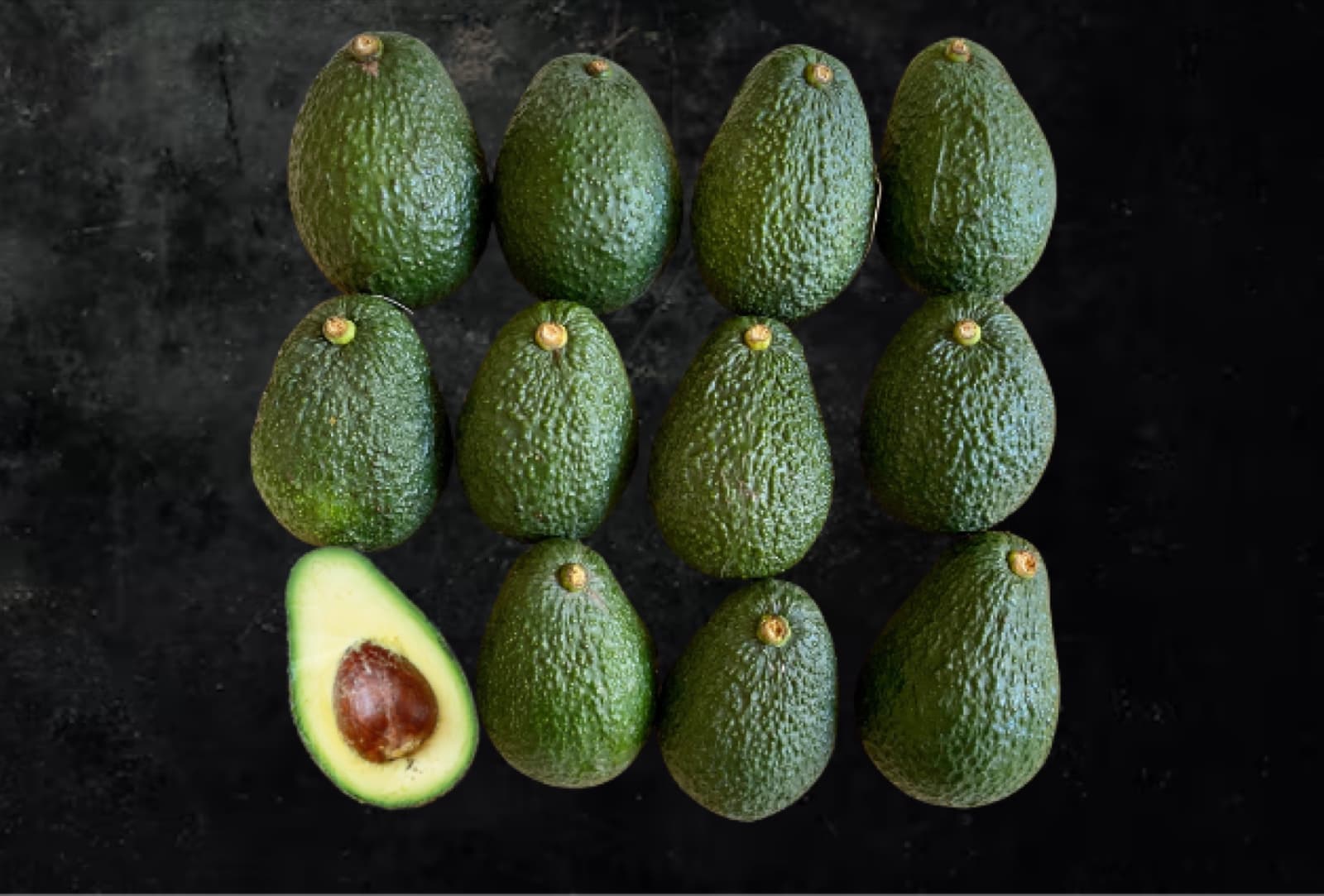 Twelve premium avocados with one cut open against a dark background